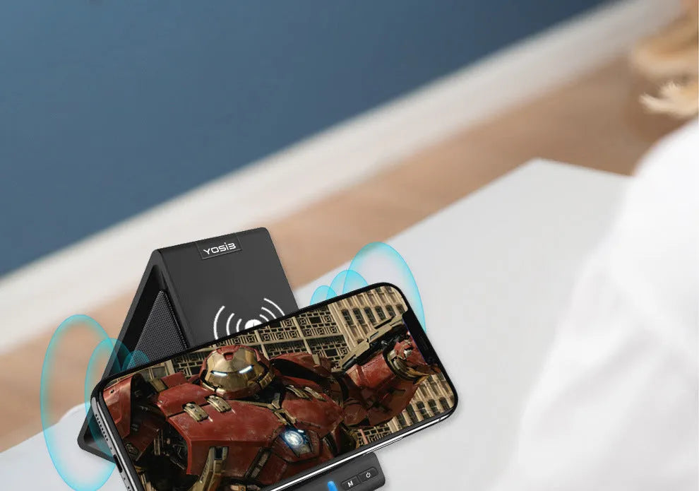 Black multifunctional Bluetooth speaker with integrated phone stand and wireless charging pad, showing size relative to a mobile phone.