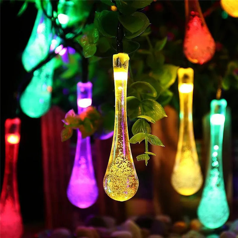 Solar-powered LED water drop string lights with bubble pendants decorating an outdoor fence at dusk.
