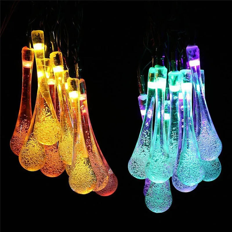 Solar-powered LED water drop string lights with bubble pendants decorating an outdoor fence at dusk.