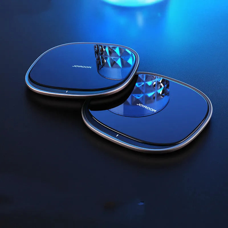 Black square 15W Qi wireless charging pad on a wooden desk next to a mobile phone.
