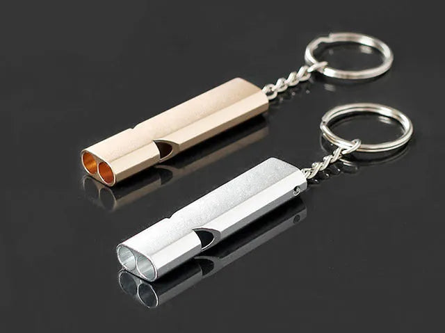 A silver or gold coloured aluminium alloy survival whistle with two holes on a grey background.