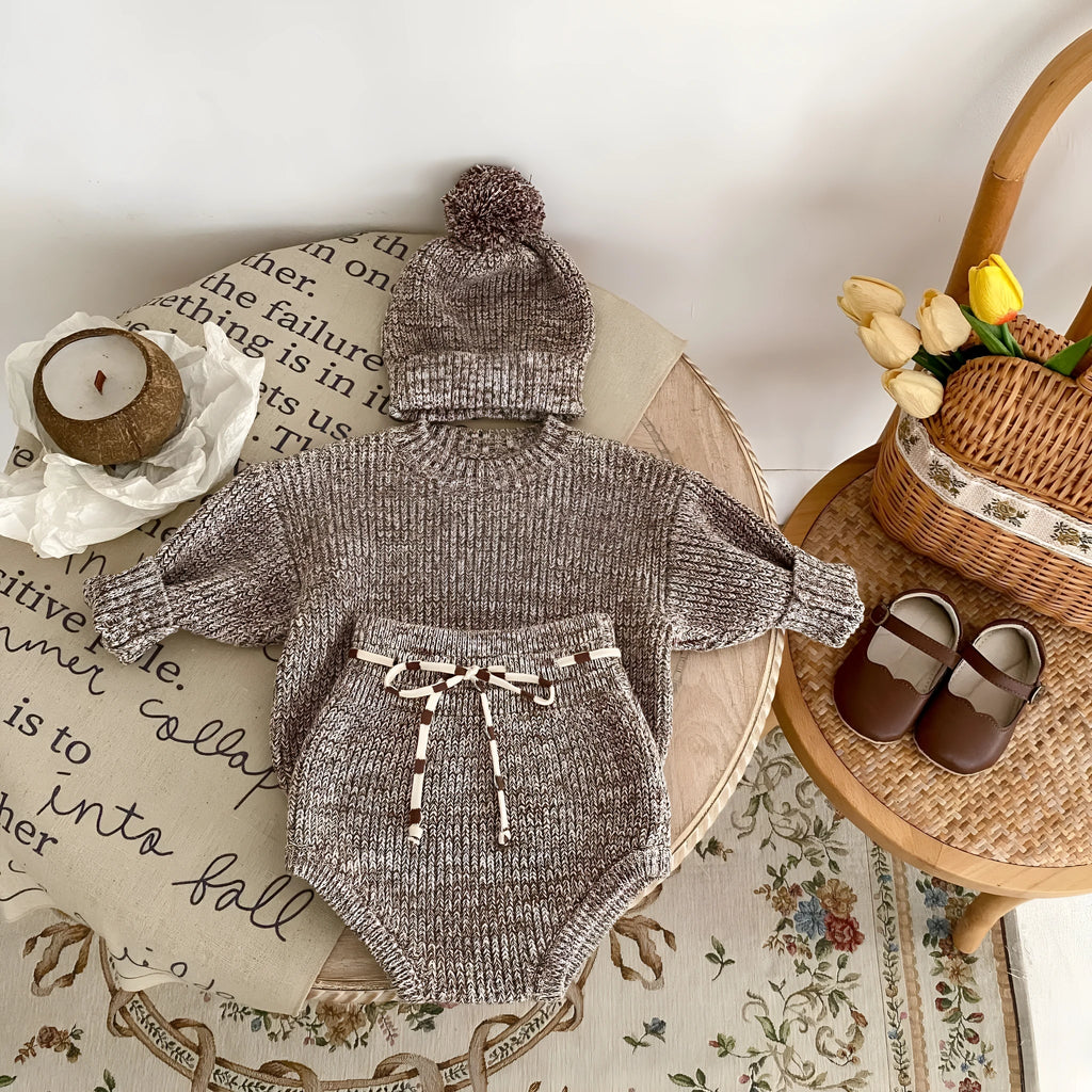 Dark brown knitted three-piece set for infant girl with fur ball hat, blouse, and shorts.