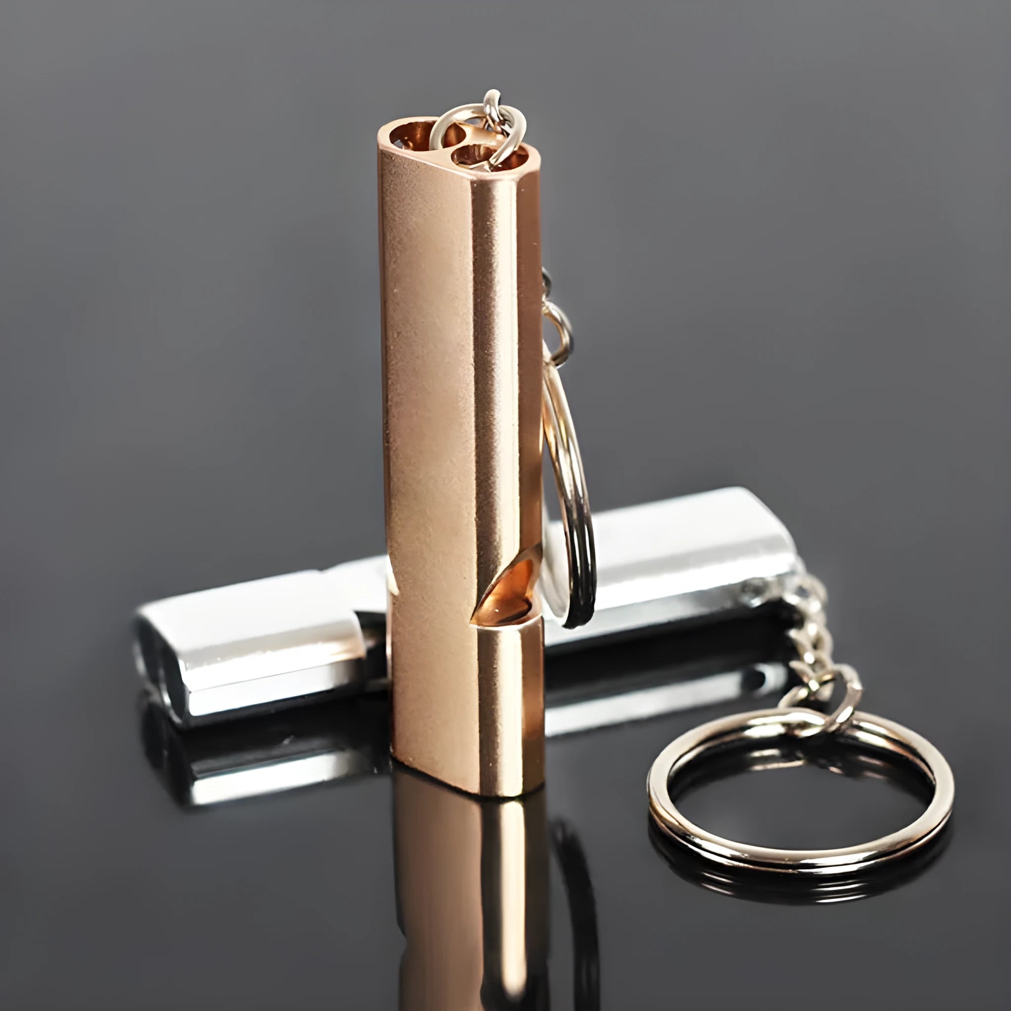 A silver or gold coloured aluminium alloy survival whistle with two holes on a grey background.