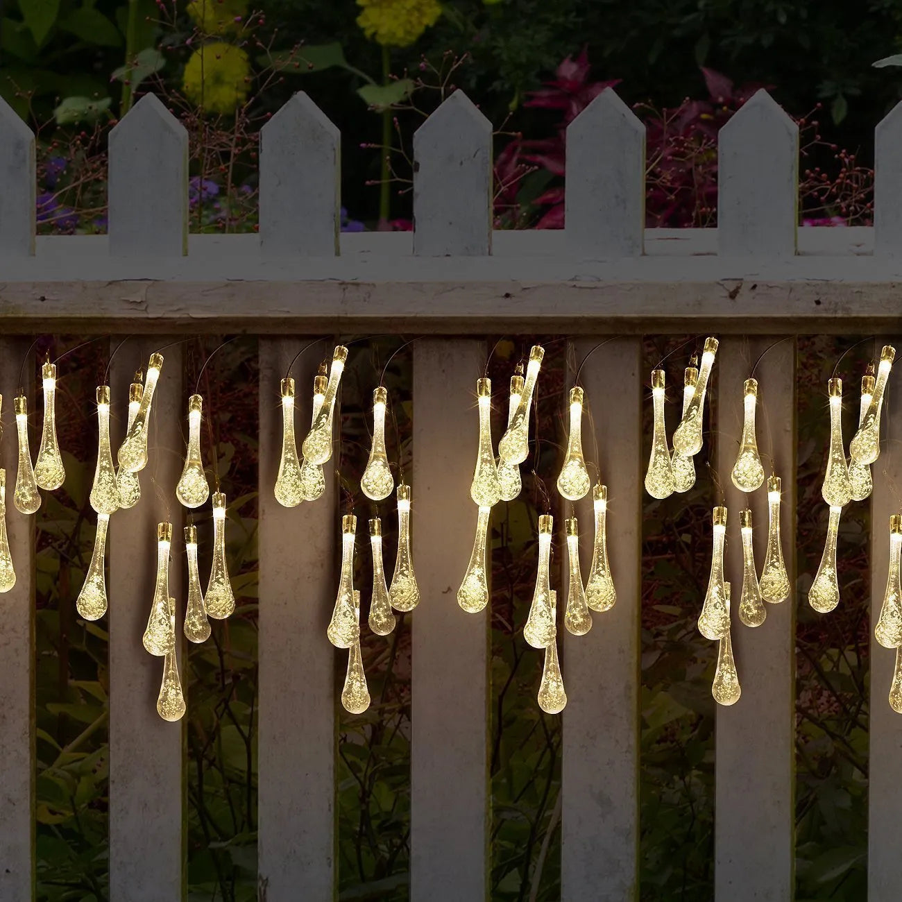 Solar-powered LED water drop string lights with bubble pendants decorating an outdoor fence at dusk.