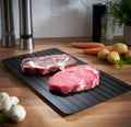 Black aluminium fast defrosting tray with silica gel feet on a kitchen countertop.