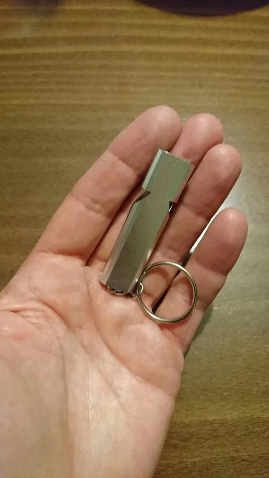 A silver or gold coloured aluminium alloy survival whistle with two holes on a grey background.