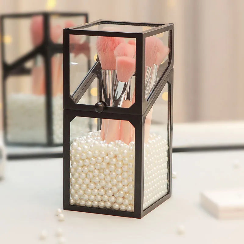 Transparent glass makeup brush bucket with black lid on white surface showing brushes inside