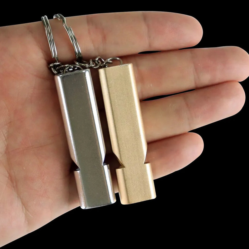 A silver or gold coloured aluminium alloy survival whistle with two holes on a grey background.