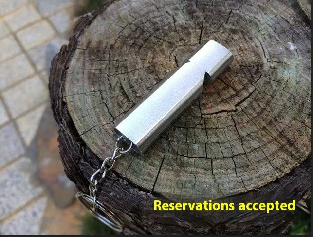 A silver or gold coloured aluminium alloy survival whistle with two holes on a grey background.