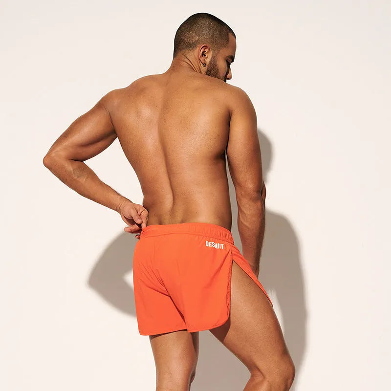 Man wearing loose orange red polyester shorts on a beach, showcasing the fit for surfing and leisure.