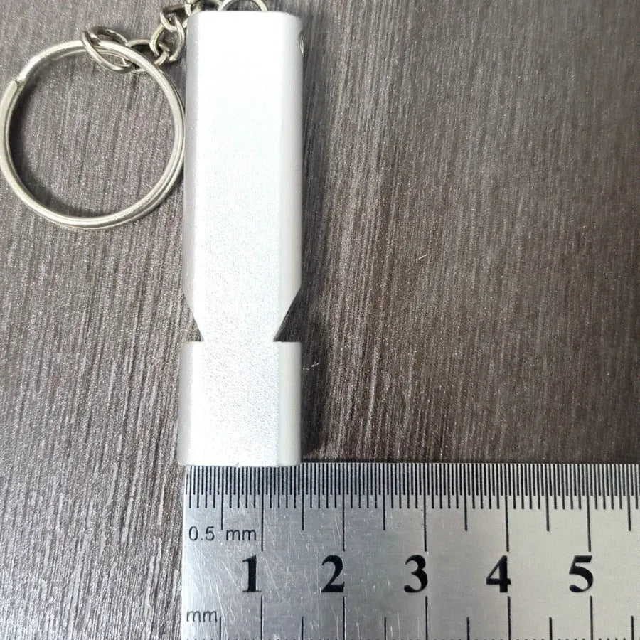 A silver or gold coloured aluminium alloy survival whistle with two holes on a grey background.