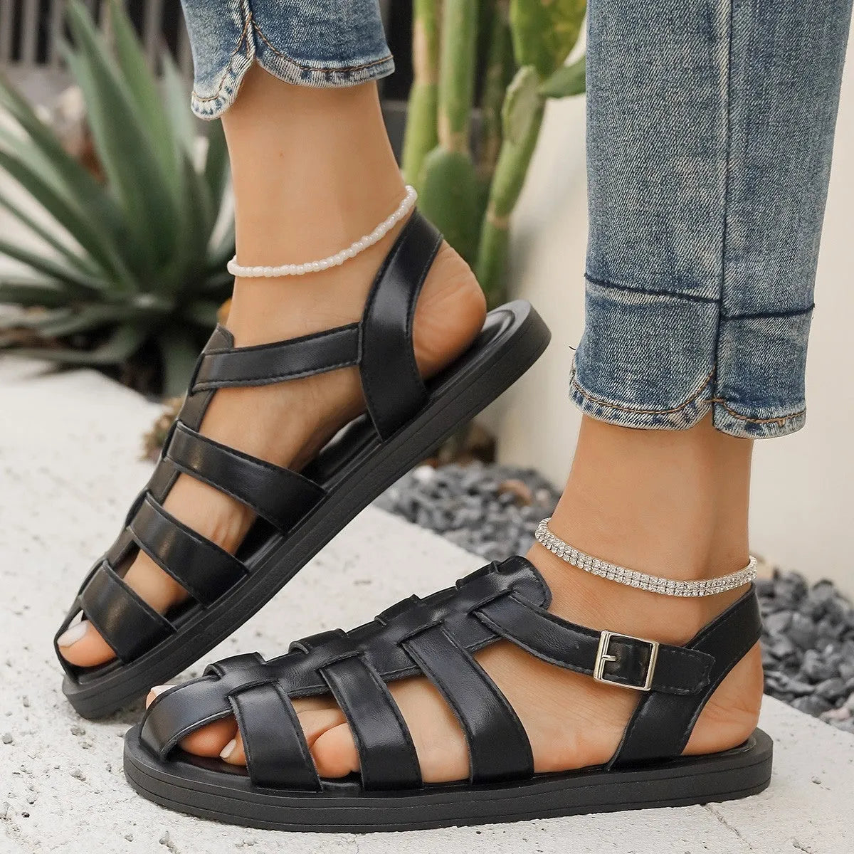 Flat Roman style women's sandals with buckle, black PU upper and rubber sole, side view