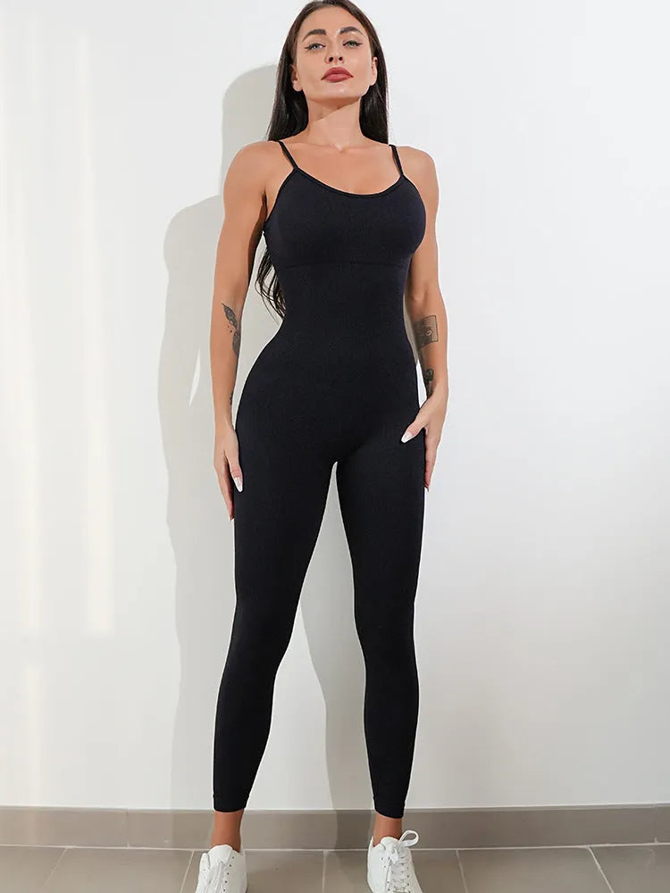 A woman wearing a black sleeveless unitard jumpsuit with spaghetti straps, showing the one-piece design and tummy control panel.