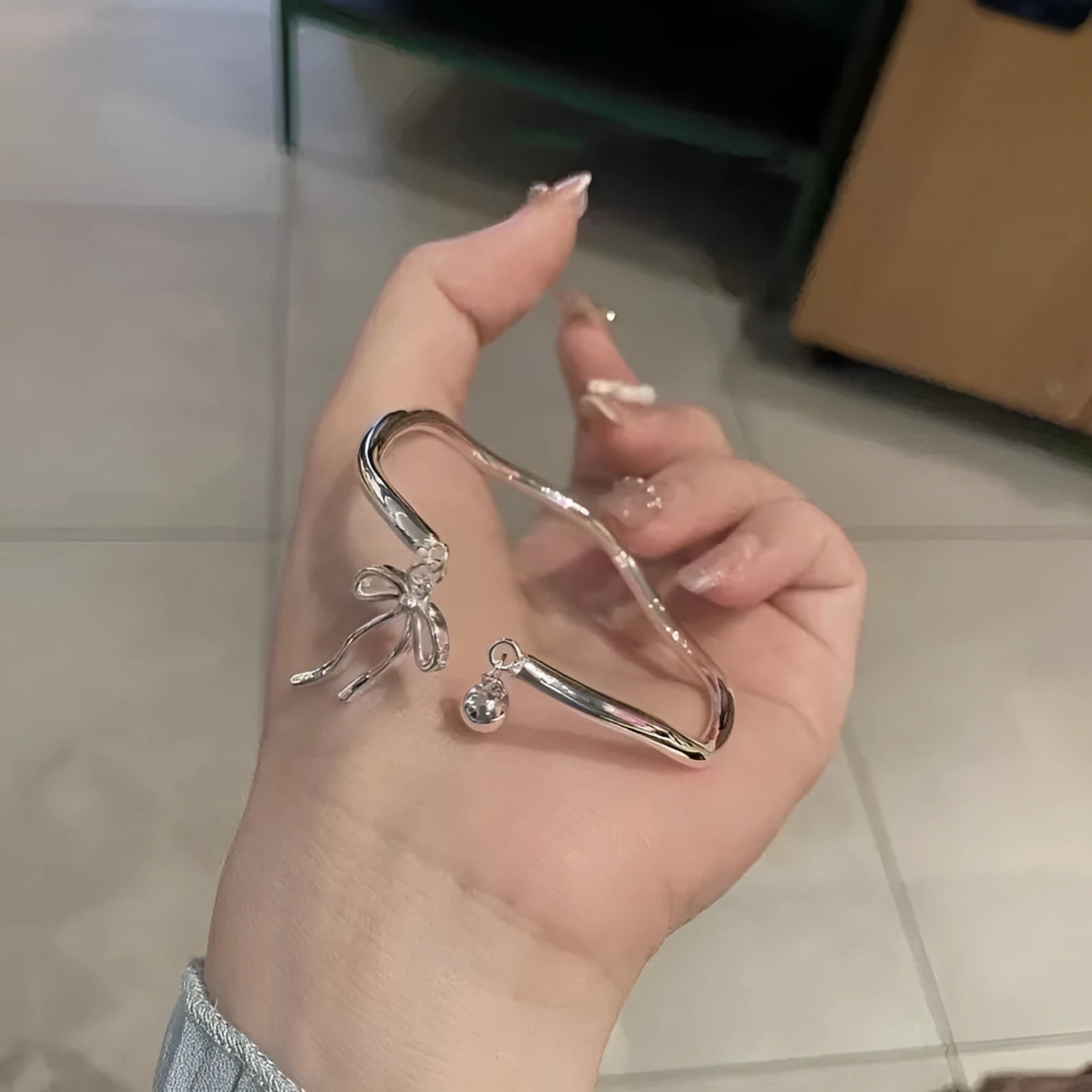 Silver adjustable bangle bracelet with a detailed bow charm and small bell on a wrist.