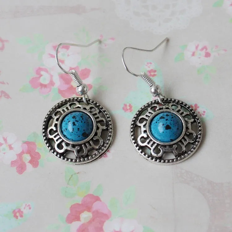 A pair of handmade ethnic earrings with a silver flower disc design, turquoise stones, and red decorative accents.