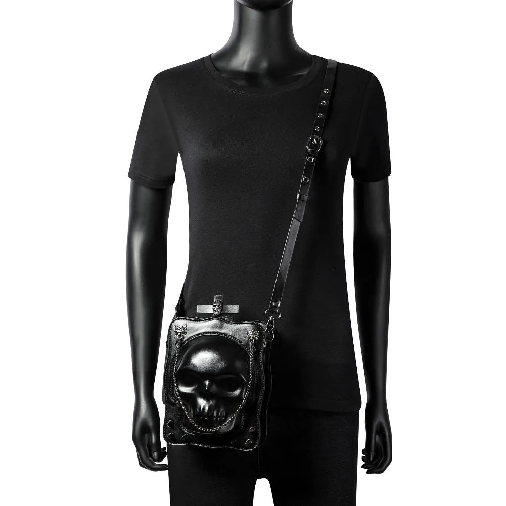 Black square crossbody bag with embossed skull design and metal chain strap against dark background