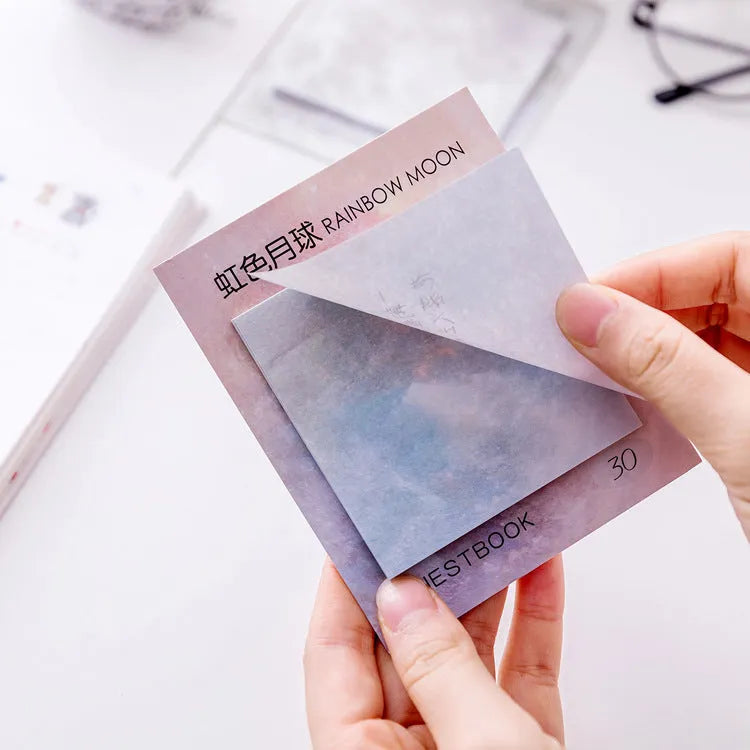 Square sticky notes notepad open to show planet oil painting designs including Earth and Rainbow Moon.